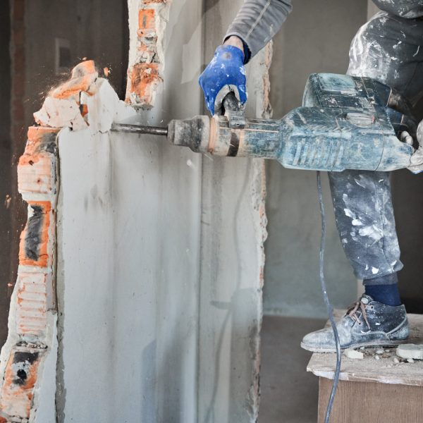 Close up view of men's hands in gloves holding electric hammer that breaking brick wall covered with plaster. Overhaul of office space, redevelopment of area.